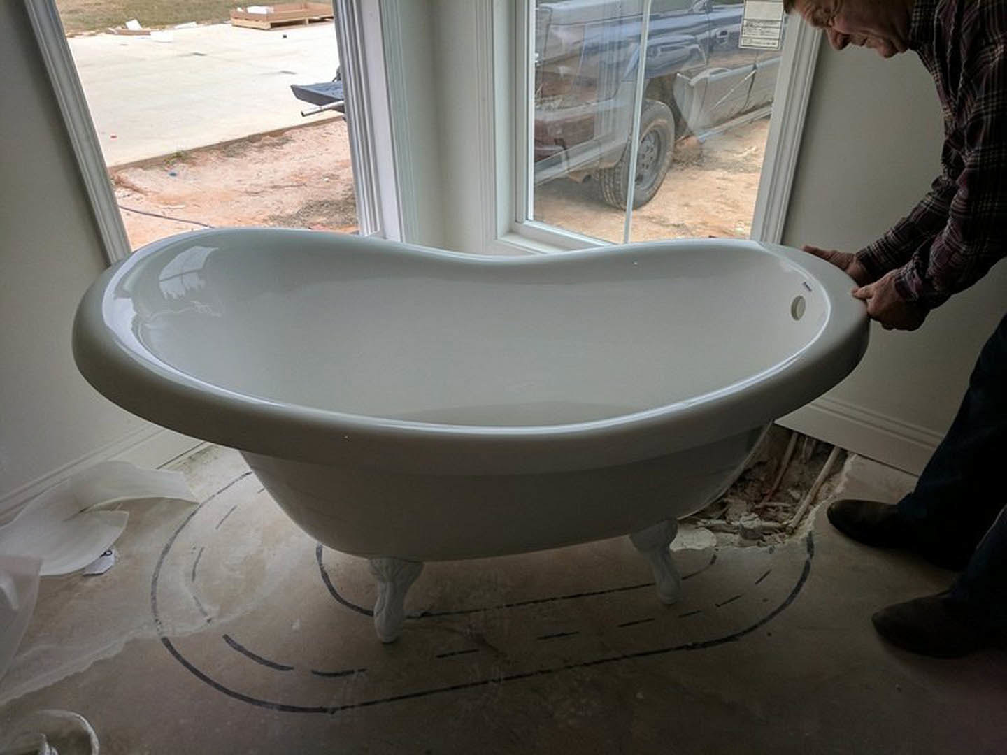 White freestanding bathtub positioned beneath a large window, man standing beside tub in modern bathroom with light-colored tile flooring and wall-mounted sink visible