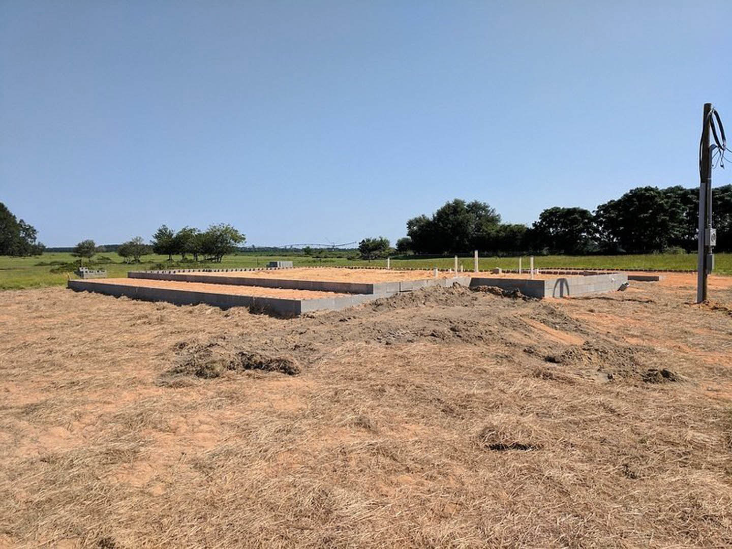 Concrete foundation set in a grassy field with patches of dirt, distant trees, and a blue sky with scattered clouds