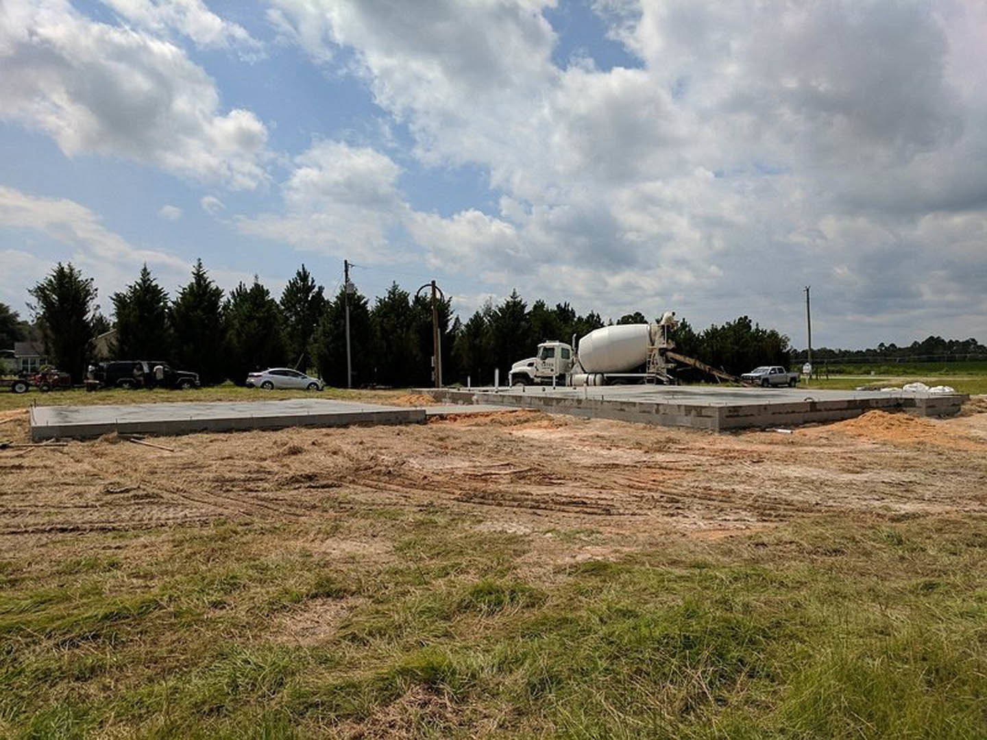 White cement mixer truck parked on a dirt patch beside a grassy field, with trees and clouds in the background, near a residential house.