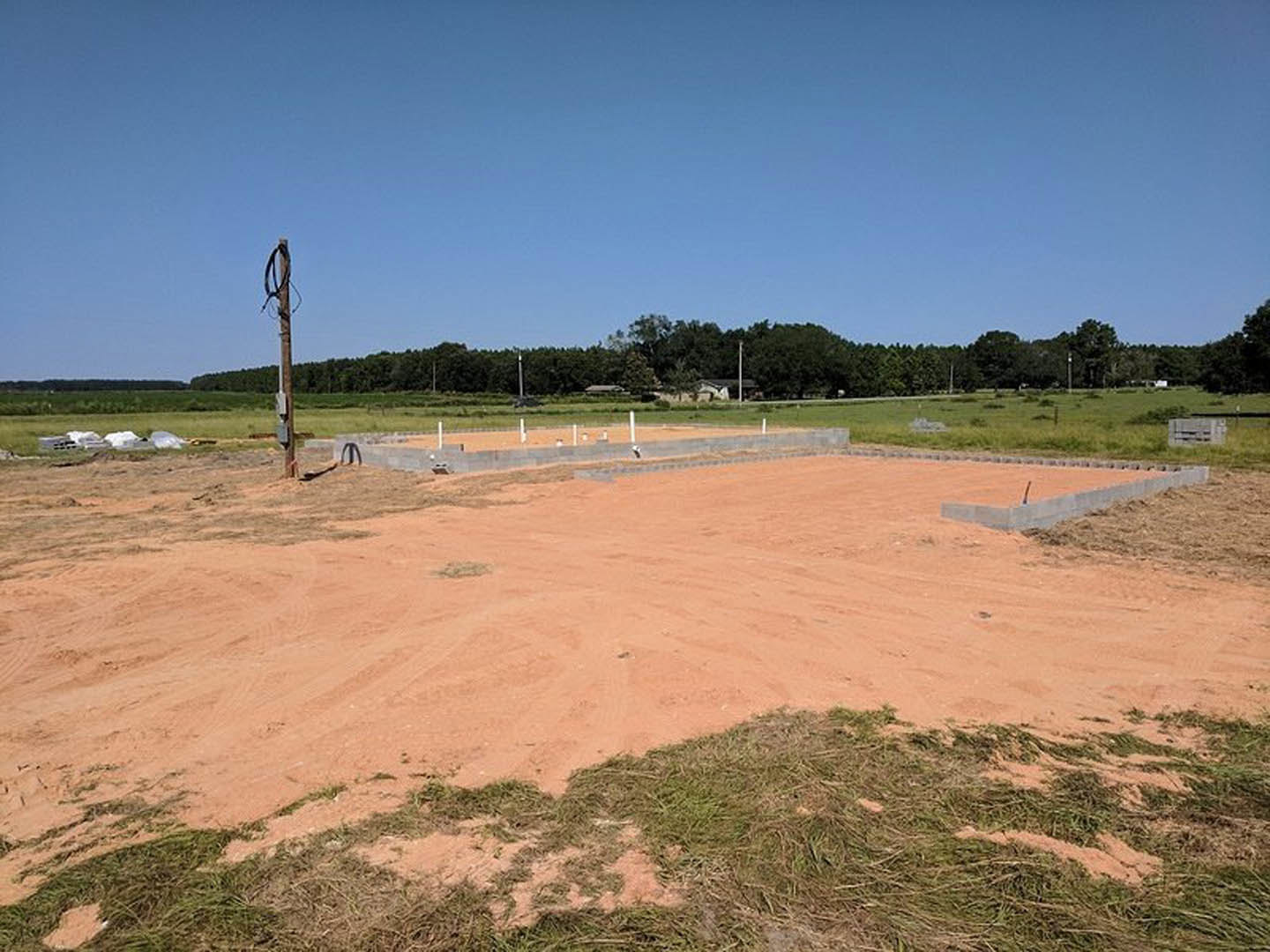 Concrete foundation slabs on a dirt construction site, surrounded by grass, trees, and a dirt road under a blue sky; light pole and fence visible in the background.