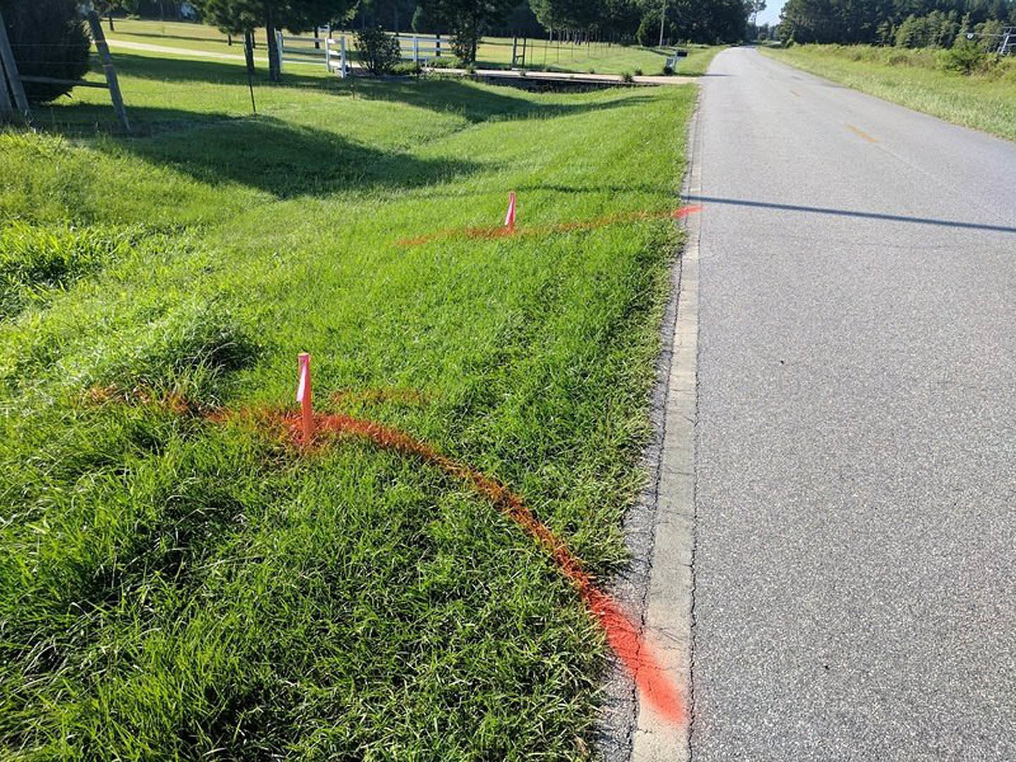 Asphalt road bordered by green grass, orange survey markers spaced along grassy edge, curb separating road from lawn, red line visible in grass, white fence in background