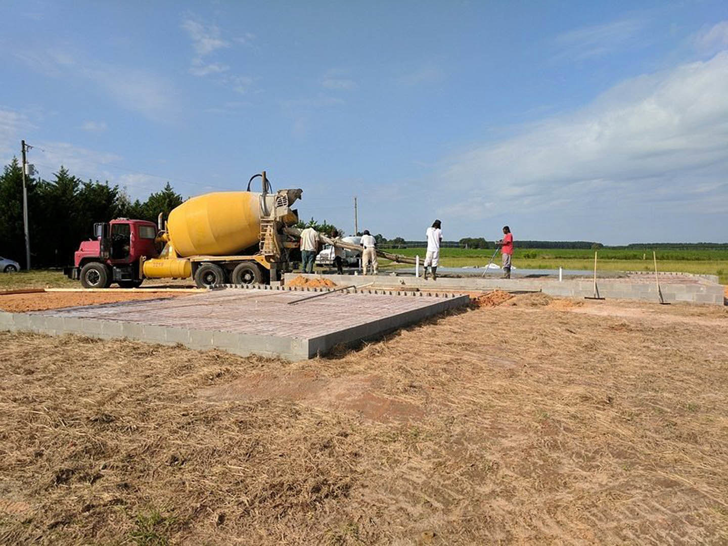 Yellow cement mixer truck parked on dirt construction site beside poured concrete slab and partially built brick wall, surrounded by grass and trees under cloudy sky