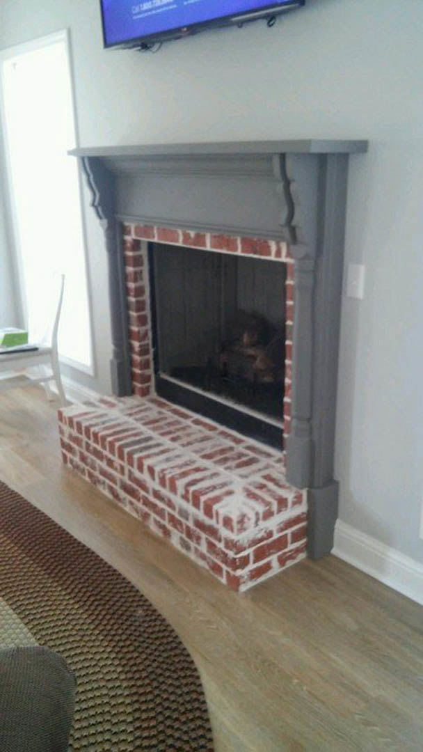 Brick fireplace with white painted surround, wood mantel shelf, fire screen, and hearth in a cozy den with hardwood flooring.