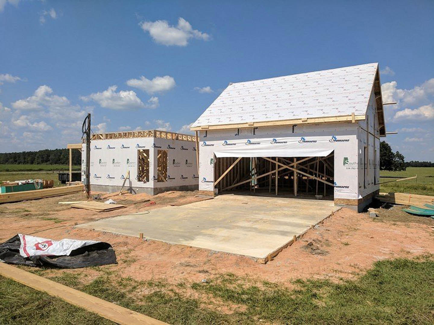 Framed house under construction with attached garage, exposed wooden beams, concrete foundation, dirt lot, white tent, and construction materials scattered around