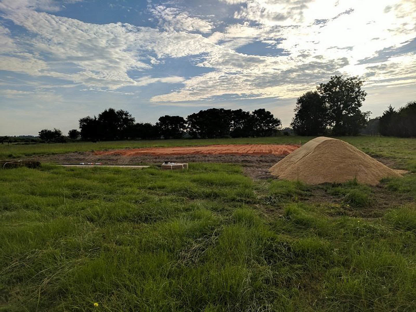 Grassy field with a large dirt mound, scattered concrete blocks, distant red hill, and trees under a partly cloudy blue sky
