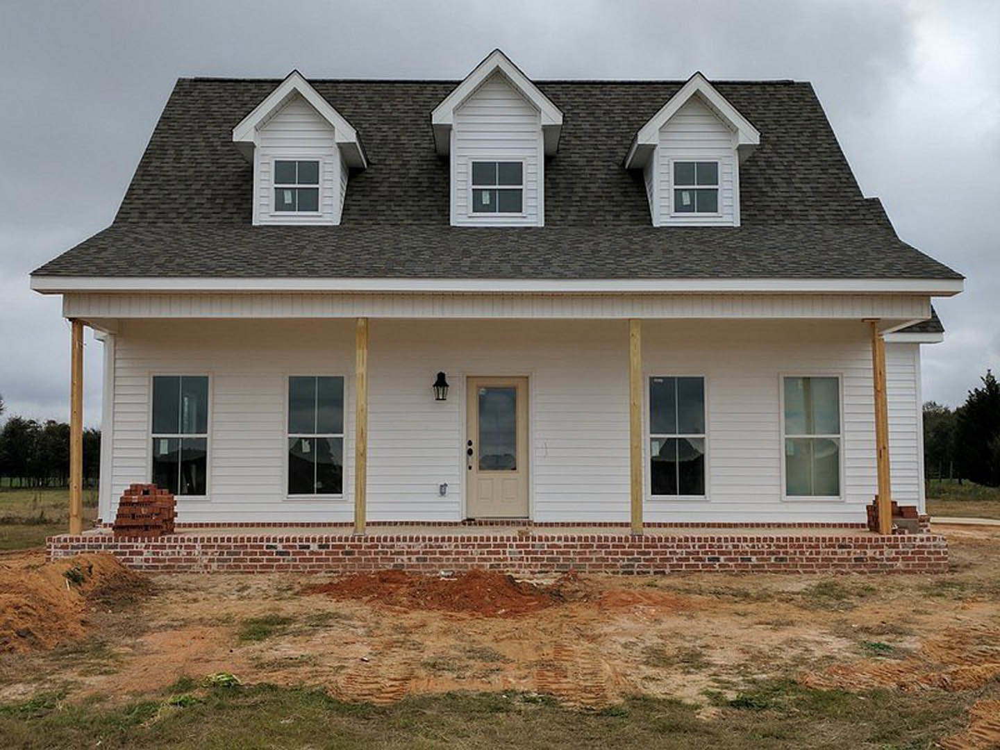 Brick house under construction with white siding, dormer windows, glass-paneled white door, and brick foundation; dirt and grass border the exterior.