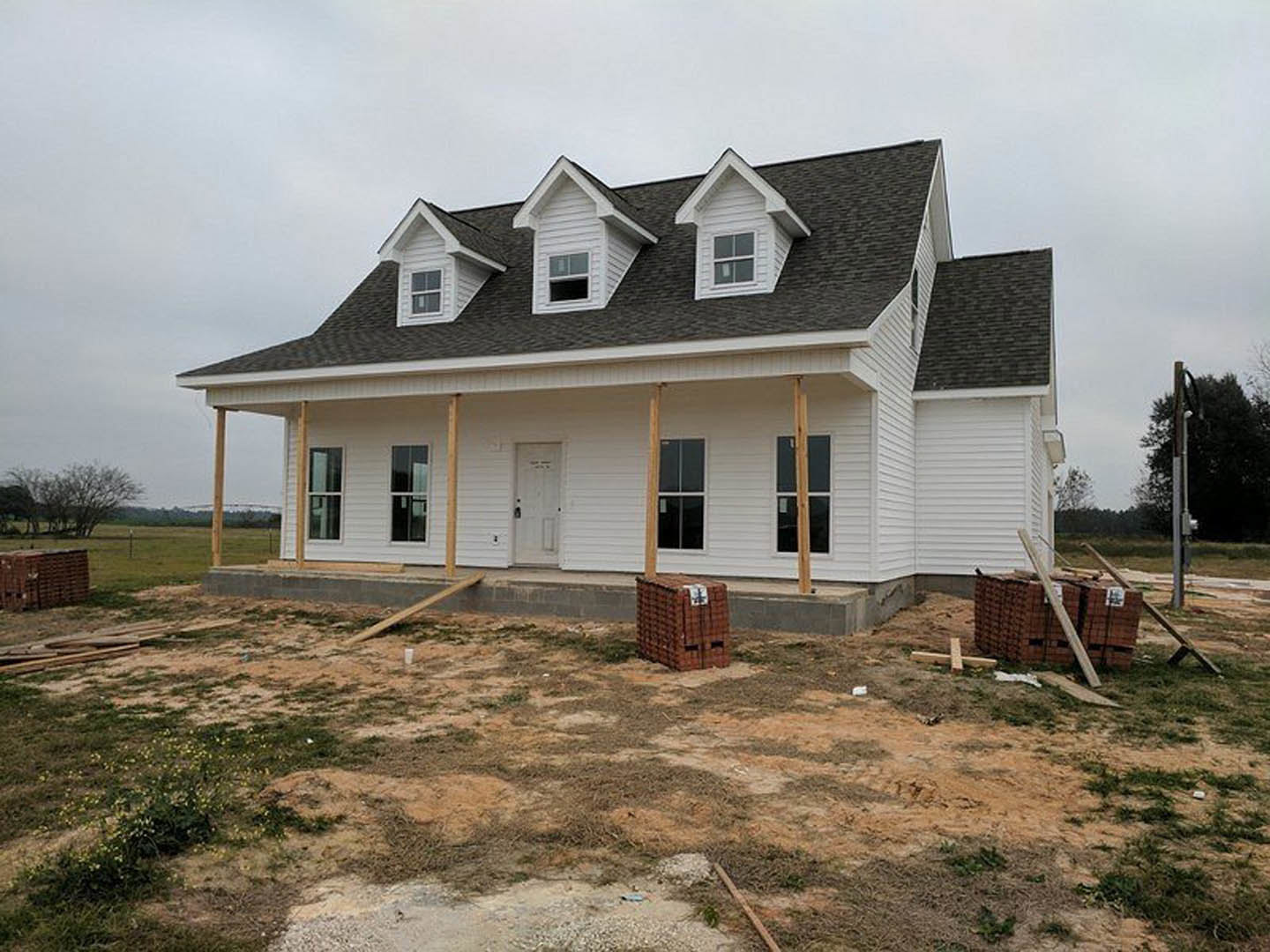 White house under construction with dormer windows, white door with black handle, stacked pallets and bricks in front, large brick box with sign, white-framed window, cloudy sky