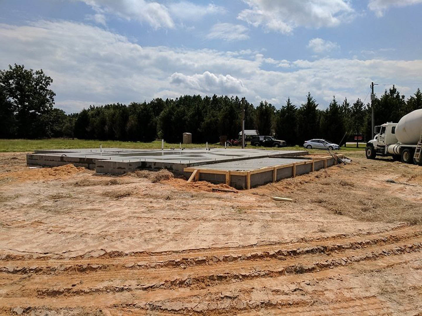 Concrete foundation with wooden beams and posts on dirt lot, large white truck parked nearby, trees and clouds in background