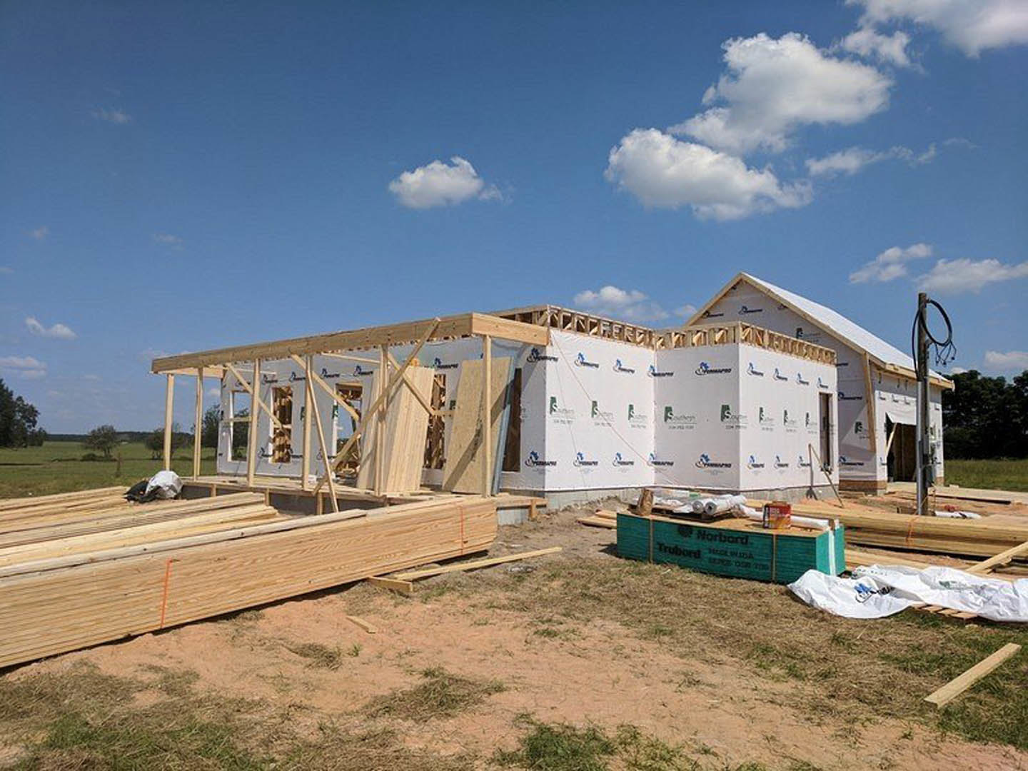 Wood-framed house under construction in open field, exposed beams and lumber piles on ground, blue sky with scattered clouds