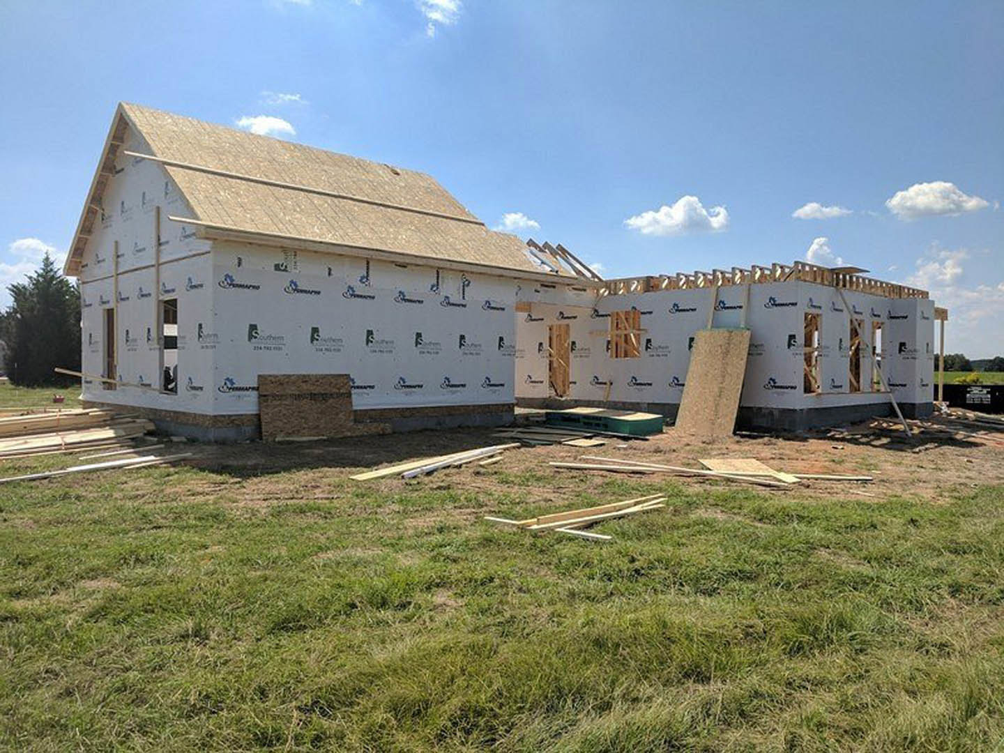 Wood-framed house under construction on grassy lot beneath blue sky, exposed beams and unfinished roof visible