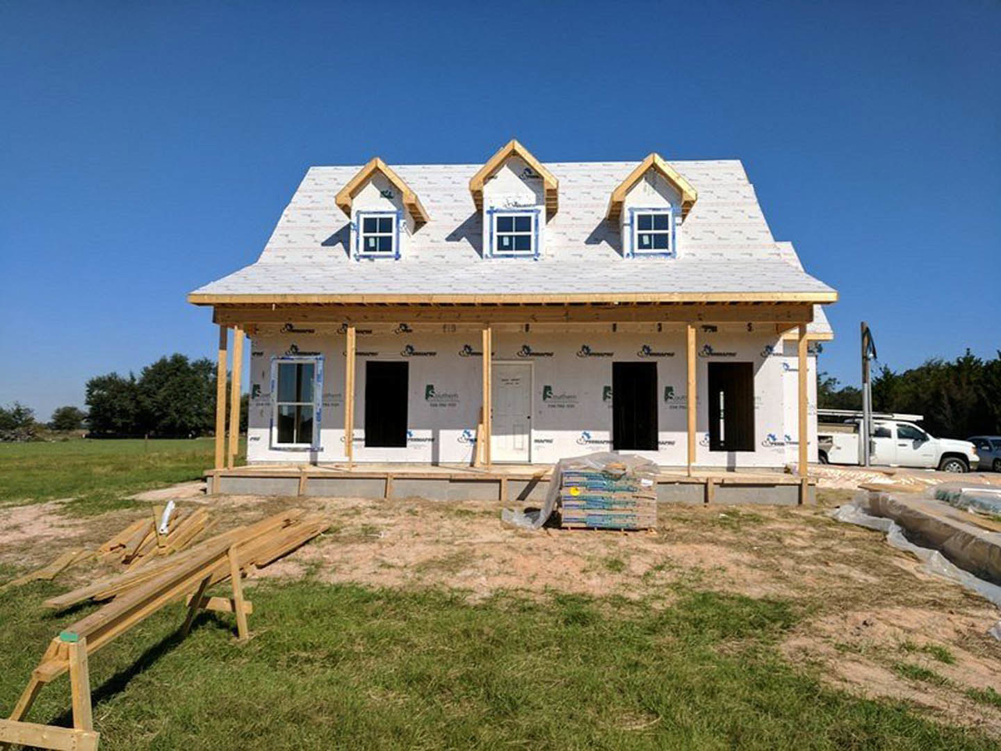 Framed house under construction in grassy field with exposed plywood walls, several installed windows, wooden planks on a sawhorse, white car parked roadside, stack of cardboard