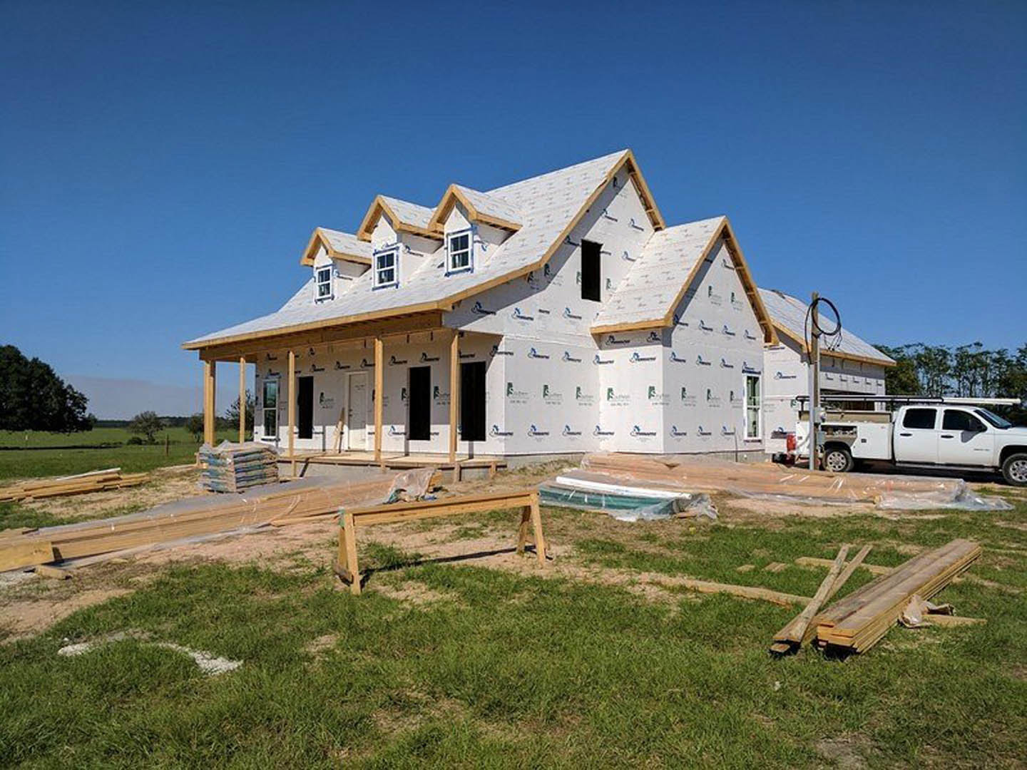 Partially built house with exposed framing, white truck parked on grass and dirt driveway, pile of lumber in foreground, blue sky overhead
