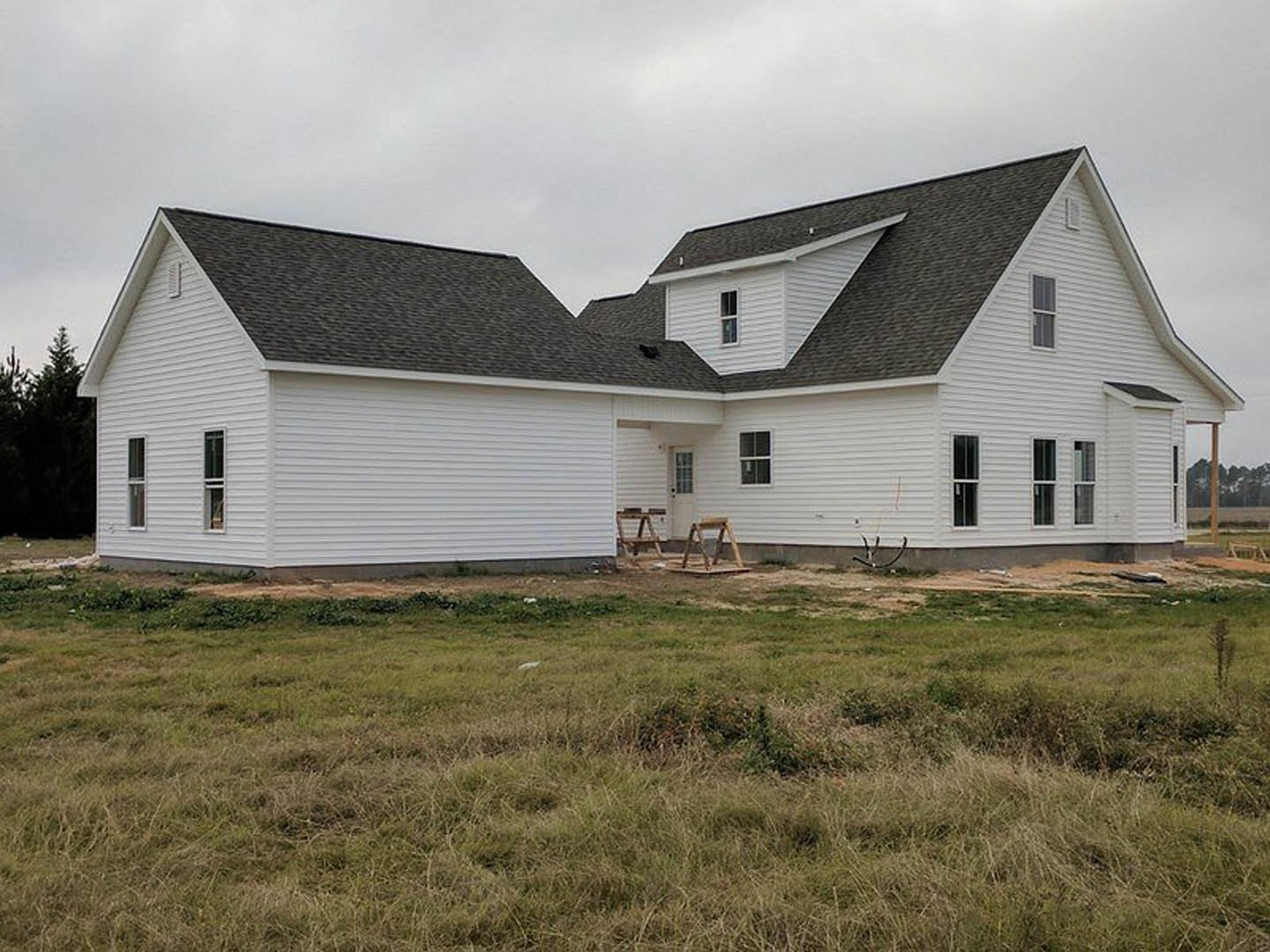 Partially built farmhouse with white siding, exposed wooden framing, garage door, wooden ladder and stool on grass lawn, cloudy sky overhead, swing set and tree in background