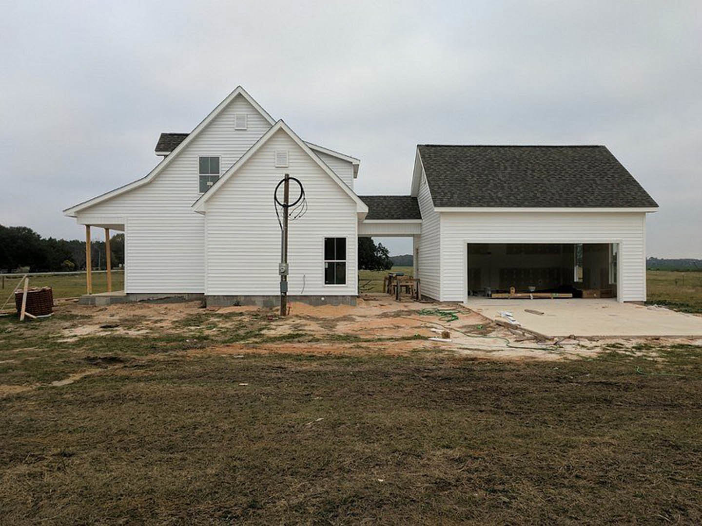 White house under construction with exposed framing, white window trim, basketball hoop in driveway, grassy yard, cloudy sky