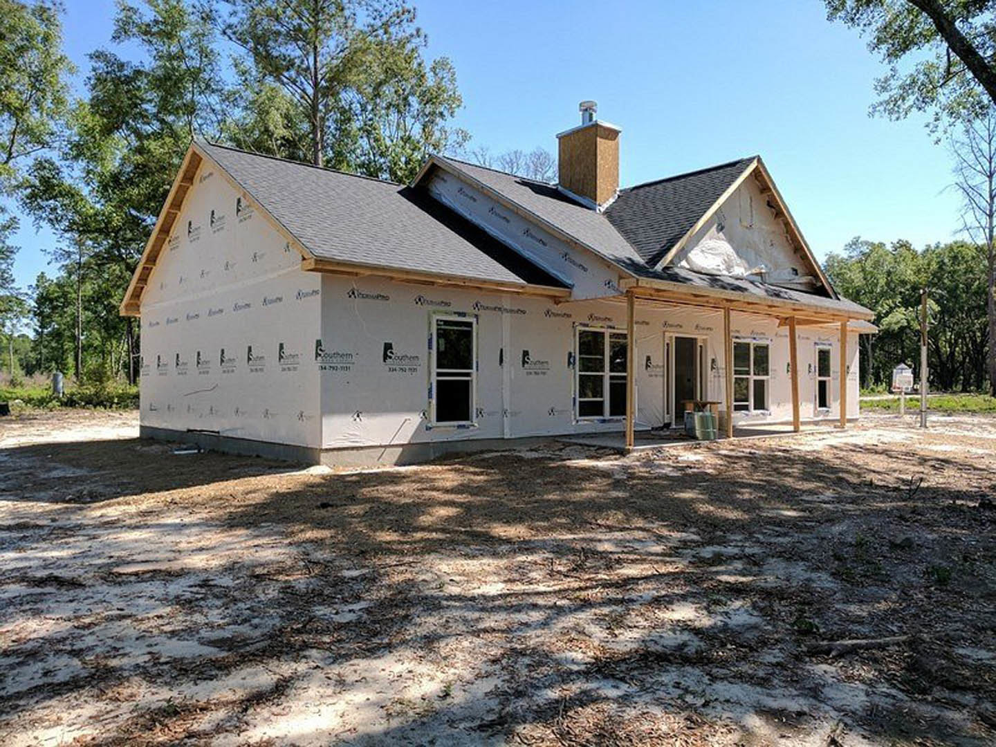 Partially built house with exposed framing, brick chimney, and unfinished windows, surrounded by trees and grassy yard, Robert Frost Farm visible in the distance