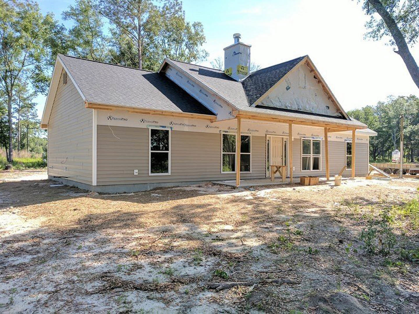 Partially built house featuring a spacious covered porch, exposed wooden framing, white window frames, and unfinished siding, set on a grassy lot with trees in the background.