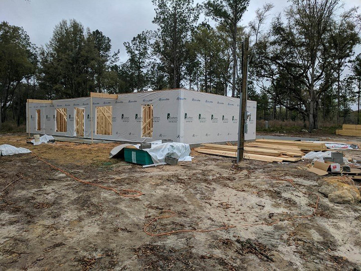 Wood-framed house under construction with exposed beams, plastic wrap, temporary fencing, and construction materials scattered on bare ground