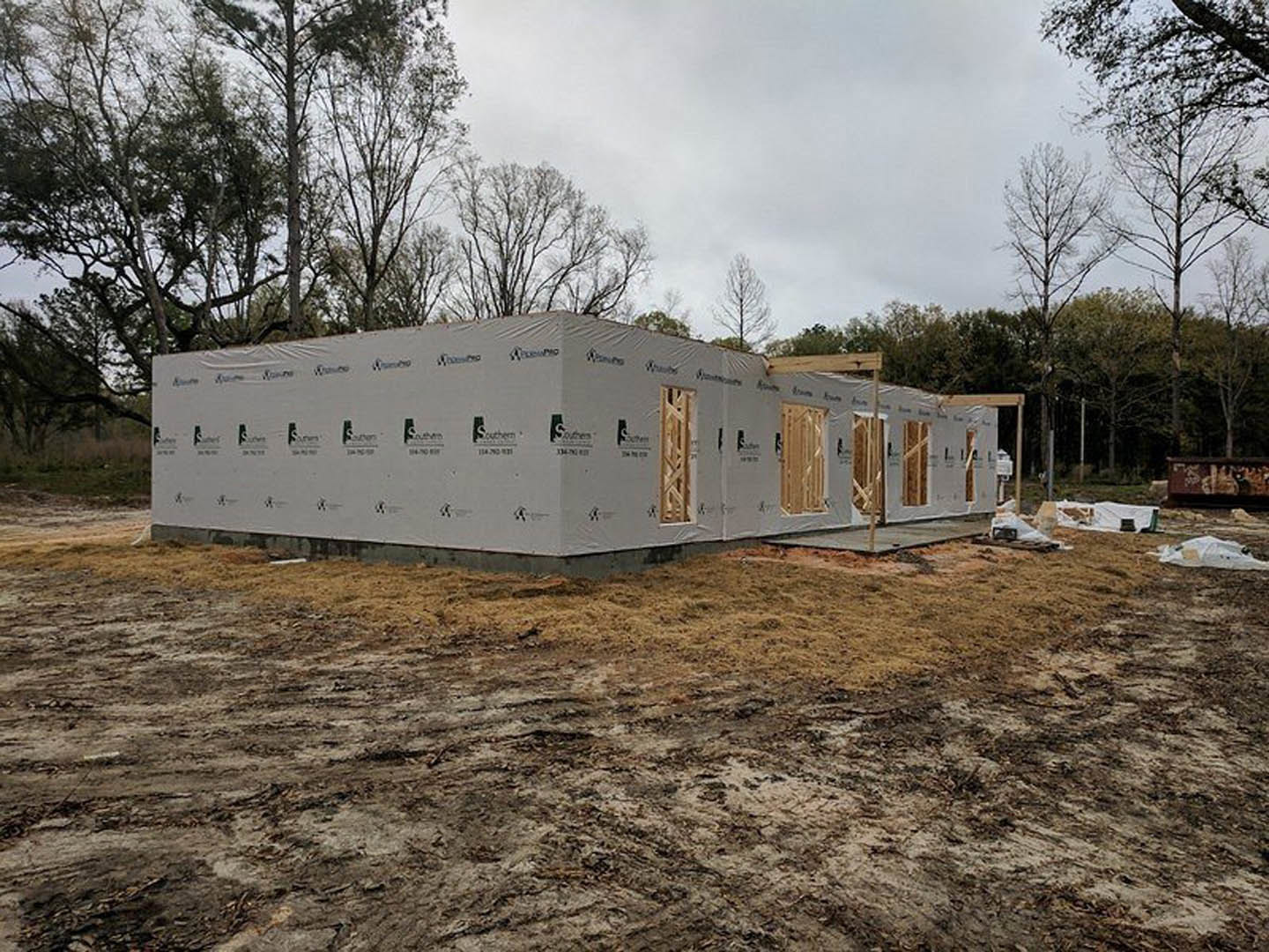Wood-framed house under construction with exposed beams, white sheeting, dirt and grass foreground, and mature trees in the background
