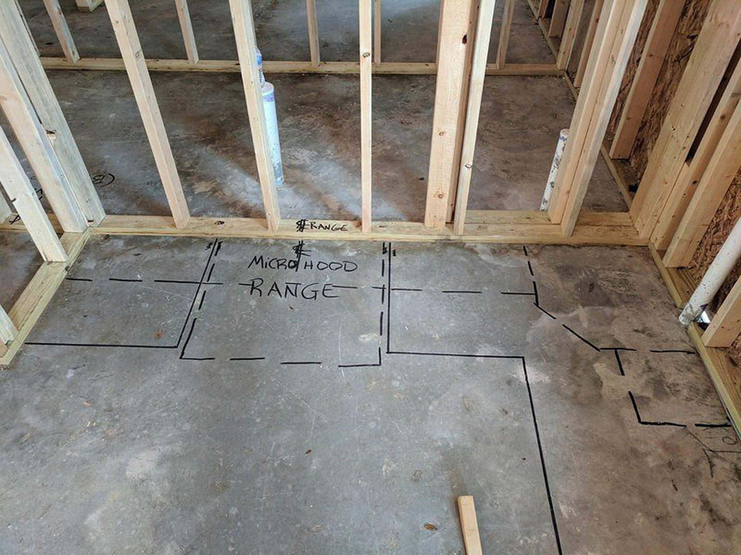 Wood-framed house under construction with black lines and architectural drawings marked on plywood floor, white support pole, partial view of construction sign, and person's leg