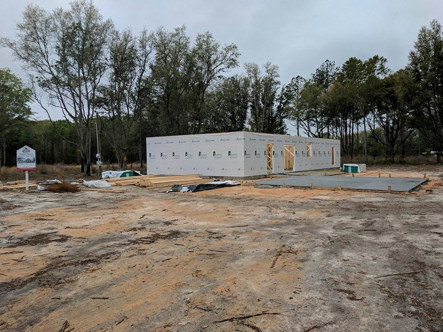 Framed custom home under construction with exposed wood beams, piles of lumber on dirt lot, trees and cloudy sky in background, white signpost near curb