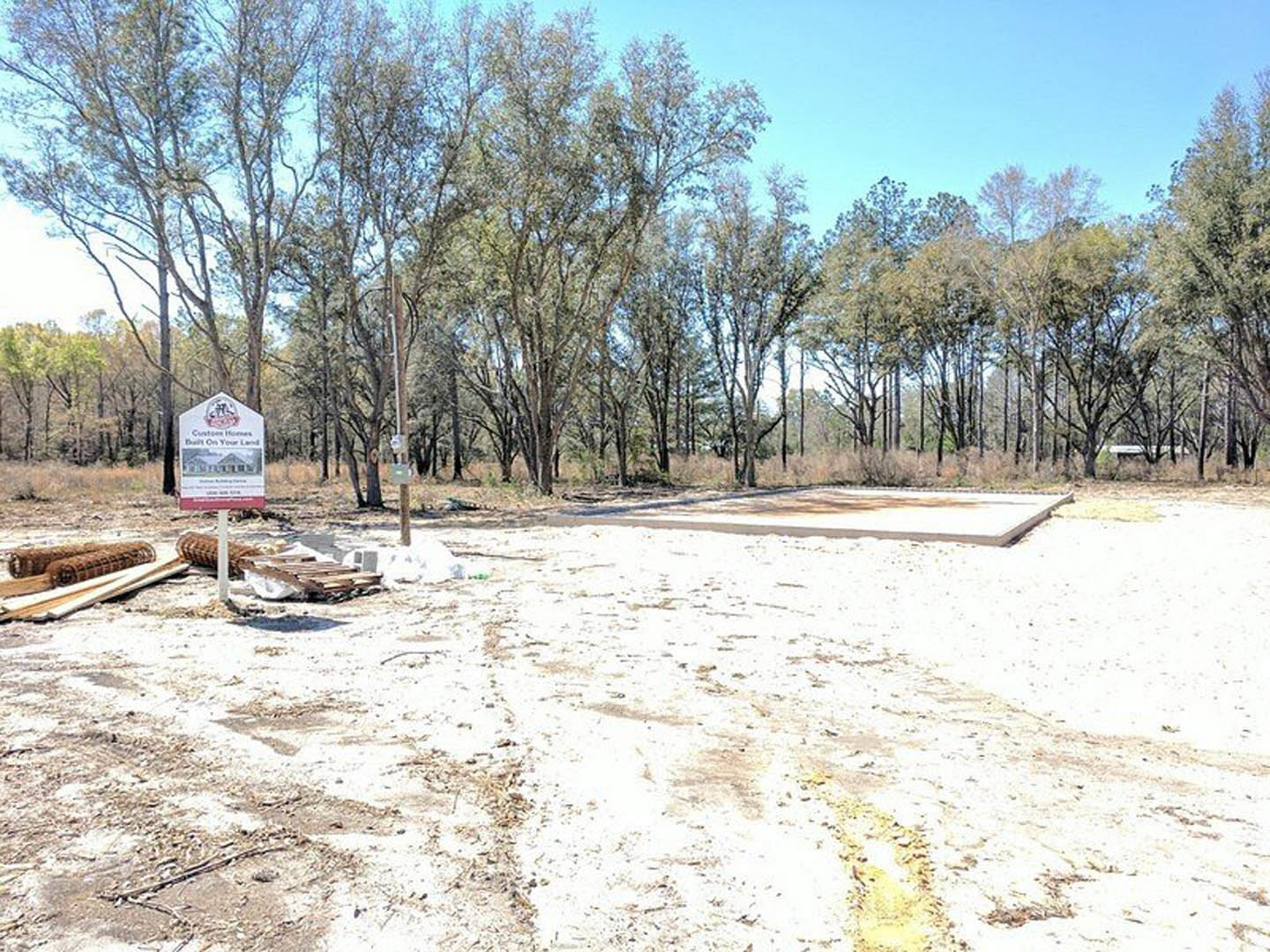 Wooden sign with a house illustration standing in the center of a dirt field, surrounded by scattered trees and open sky in the background