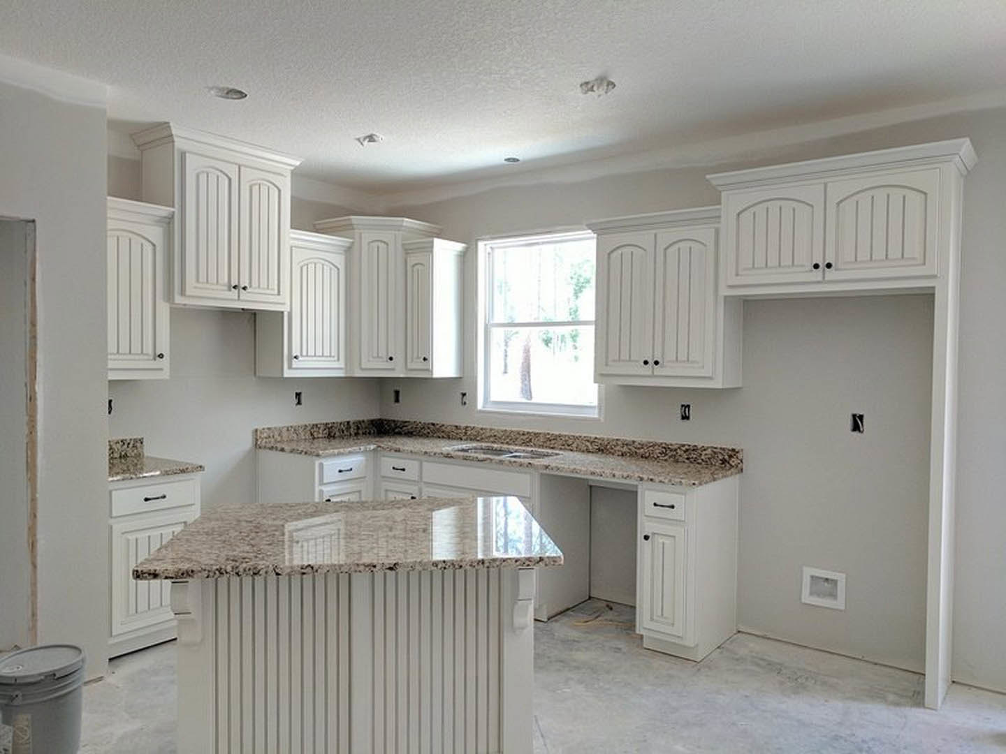 White kitchen cabinets with marble countertop, white walls, window with white frame, close-up of a bucket, white drawer with metal handle, and white square frame with hole.