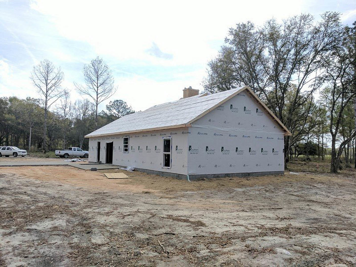 Two-story house under construction with exposed framing, partially finished roof, dirt lot scattered with debris, pickup truck parked in front, white car on nearby road, trees and