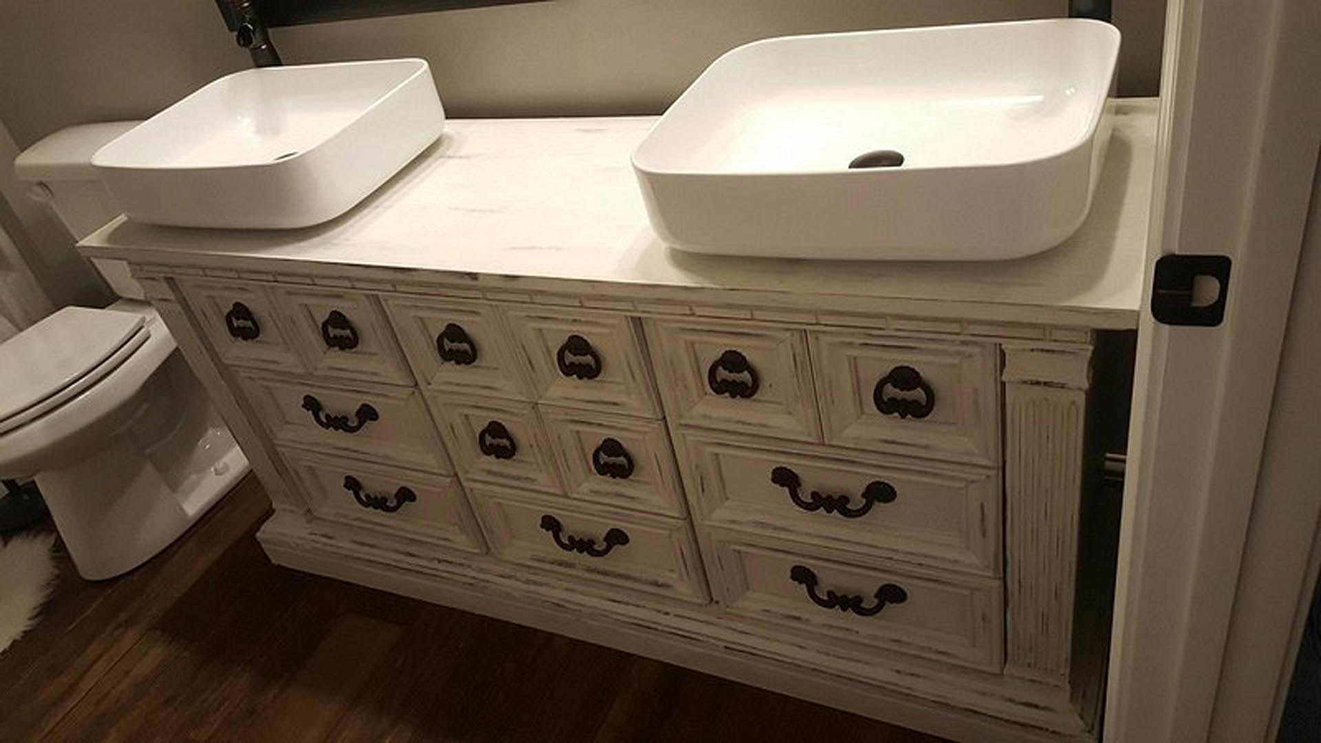 Double white vessel sinks with chrome faucets set atop a white dresser-style vanity, surrounded by light tile walls and cabinetry in a modern bathroom.