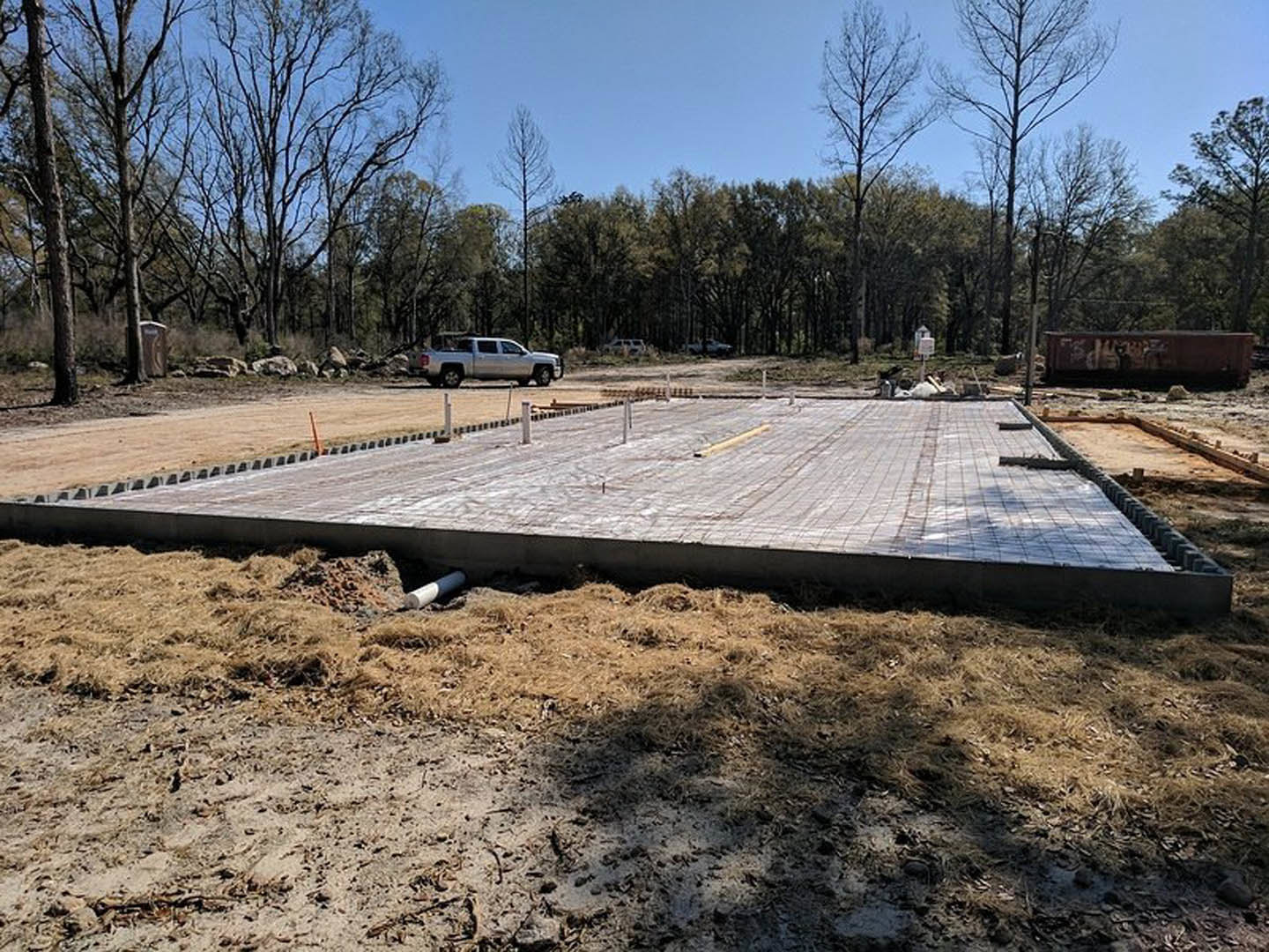 Freshly poured concrete slab with metal poles, white truck parked on dirt road, white pipe in soil, trees and plants in background, cat perched on wall.