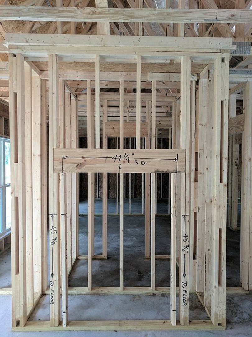 Exposed wooden framing and beams inside a partially constructed house, set on a concrete floor with window openings visible