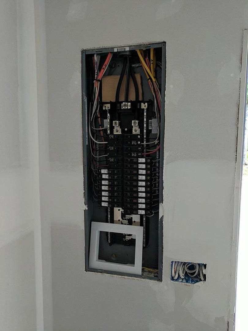 Electrical panel mounted on interior wall with exposed wiring and cables, black metal cover and white label visible