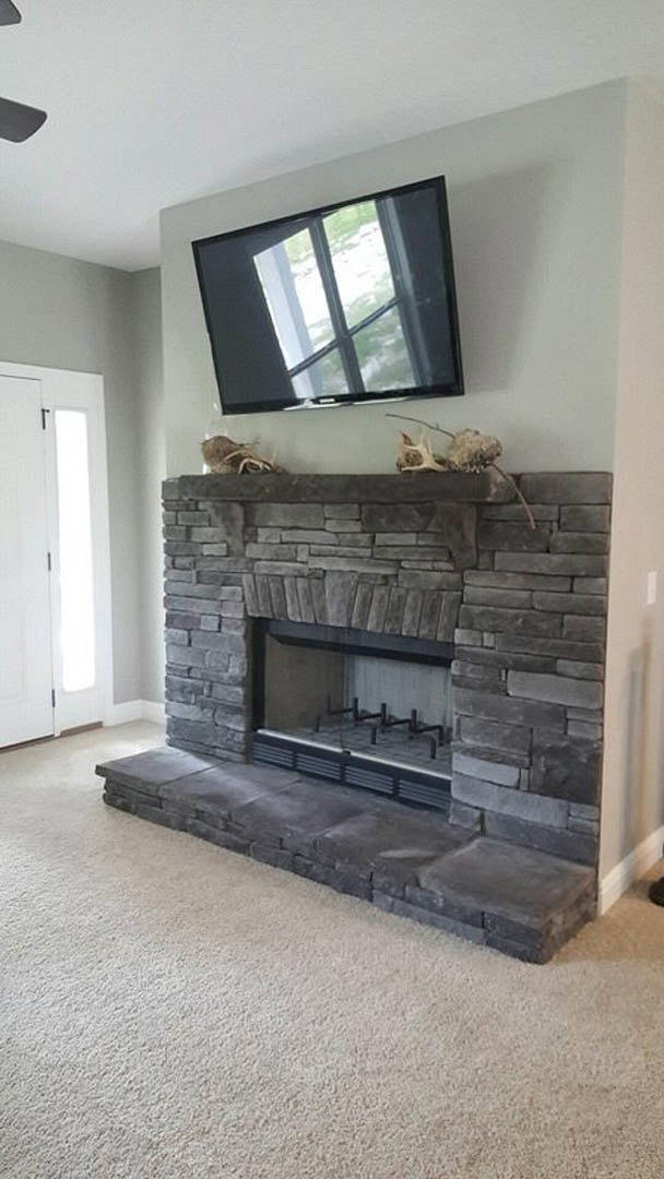 Stone fireplace with flat screen TV mounted above, white carpet flooring, stone mantle, white door with sunlight streaming through, television screen reflecting nearby window