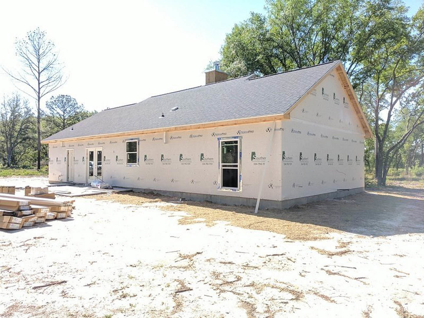 Partially built house with exposed framing, white exterior walls, large window with white frame, leafless tree nearby, construction materials scattered on bare ground