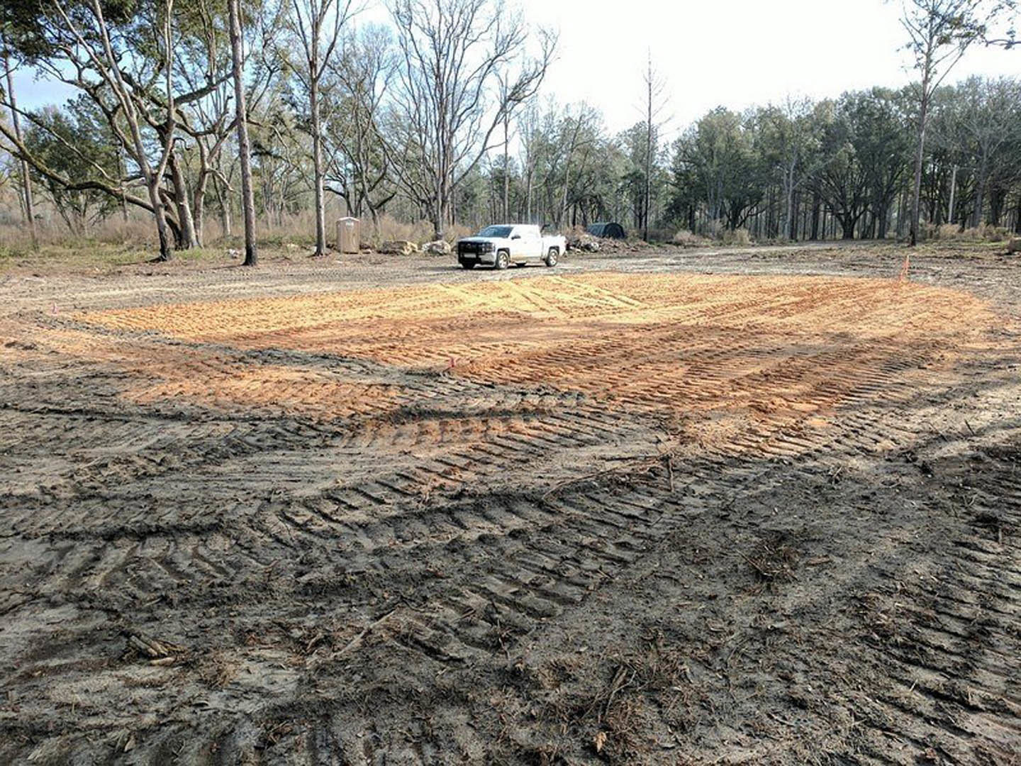 White pickup truck parked on a dirt driveway beside a custom home, surrounded by tall trees and grassy field under an overcast sky