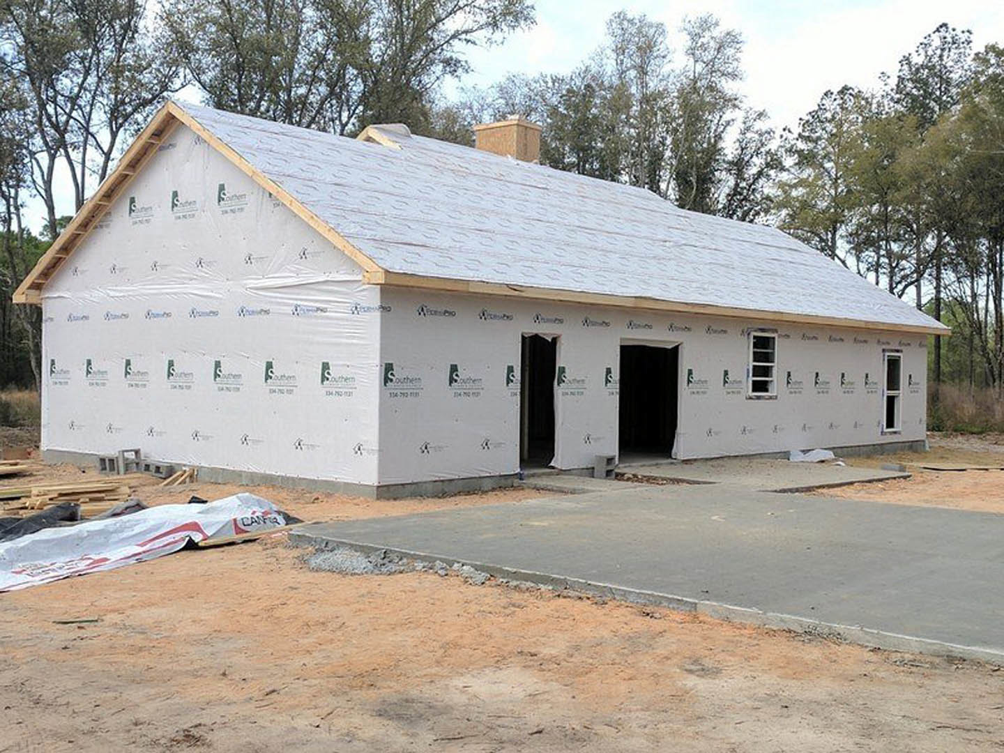Partially built house surrounded by exposed dirt, white and red tarps covering ground, person in black suit near structure, white-framed window visible, trees and sky in background