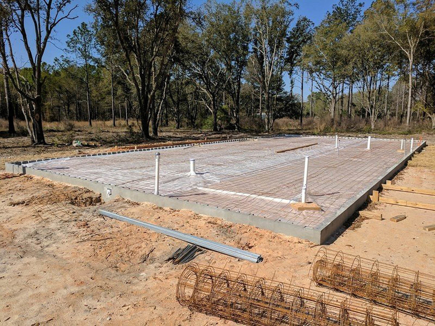 Concrete foundation slab with white poles and wire mesh, surrounded by sandy soil and metal rods, forest trees in the background.