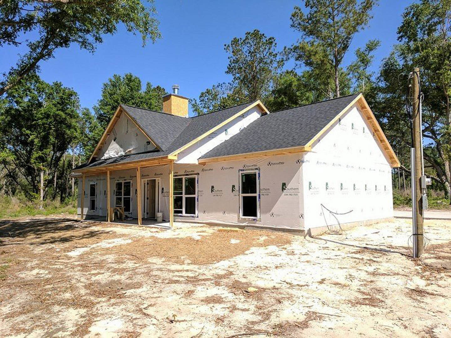 Framed house under construction with white sheathing, exposed wooden beams, several installed windows, chimney, and surrounding trees in the background