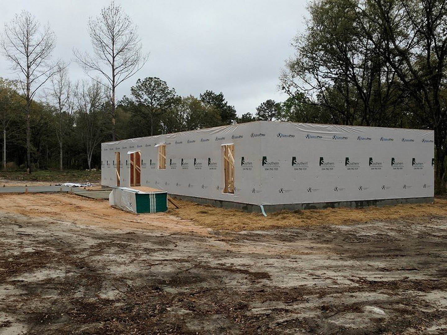 Partially built white exterior walls with open doorway, exposed construction materials, leafless tree in foreground, clear sky overhead