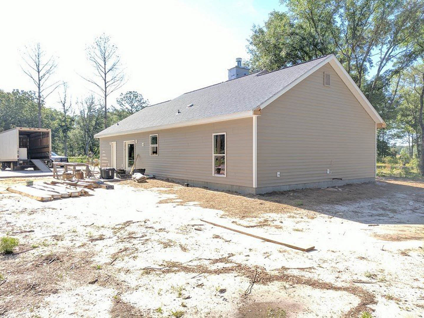 Partially built house with exposed wooden framing and white window trim, surrounded by tall trees; construction truck parked nearby on dirt patch, small green plant in foreground