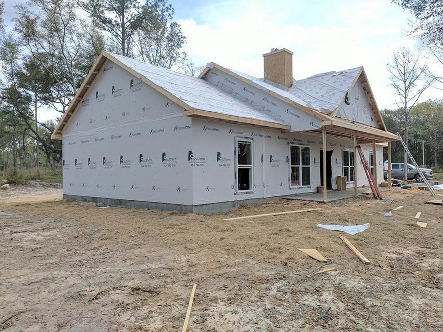 Wood-framed house under construction with exposed insulation, white-framed windows, ladder leaning against exterior, parked car beside utility pole, trees and sky in background