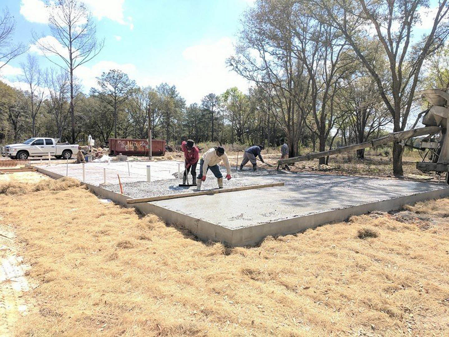 Workers preparing and leveling a concrete slab foundation outdoors, surrounded by grass, trees, and construction materials, with a white car parked nearby