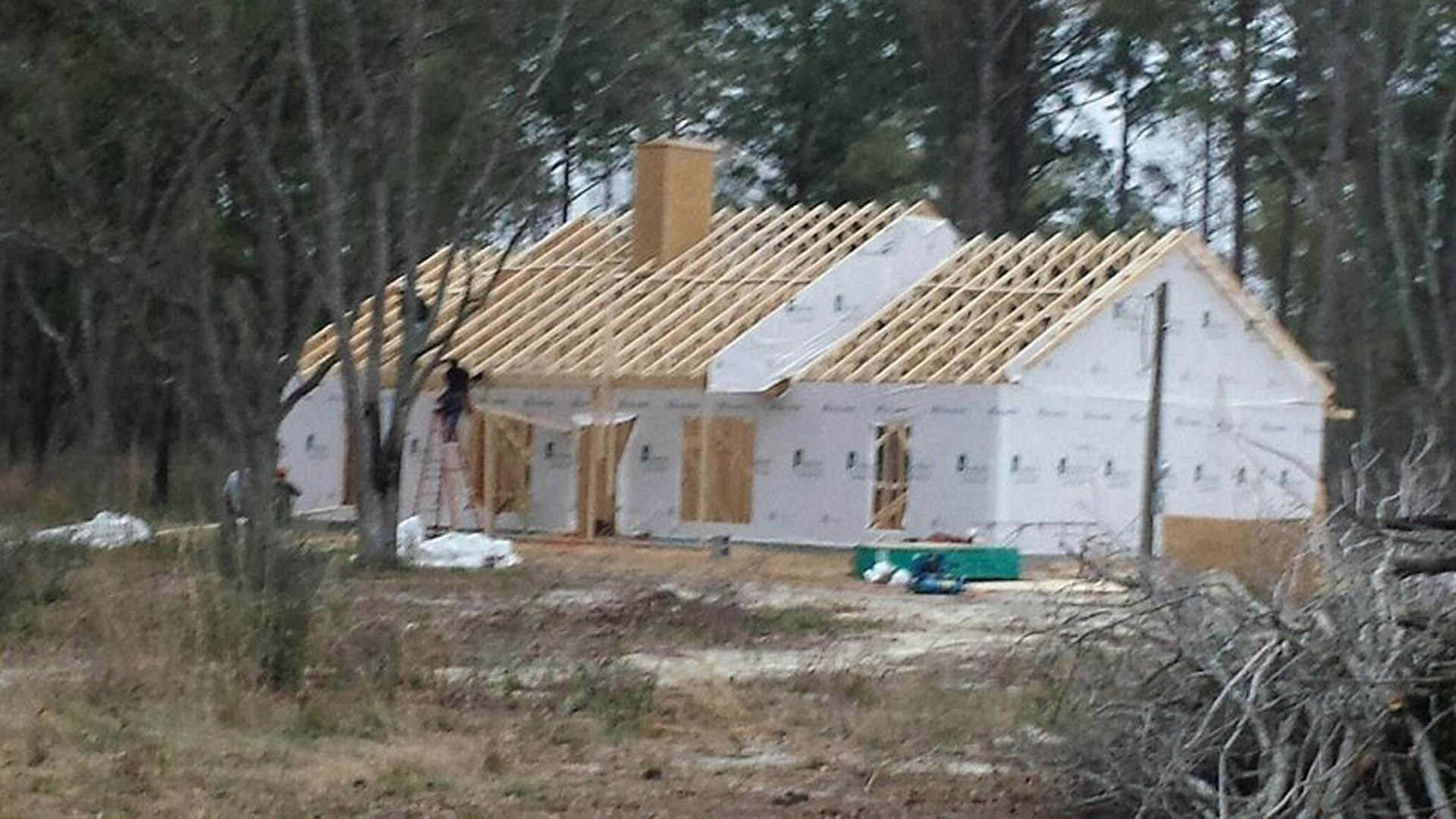 Wood-framed house under construction with exposed beams, ladder leaning against unfinished exterior, grassy yard, and surrounding trees