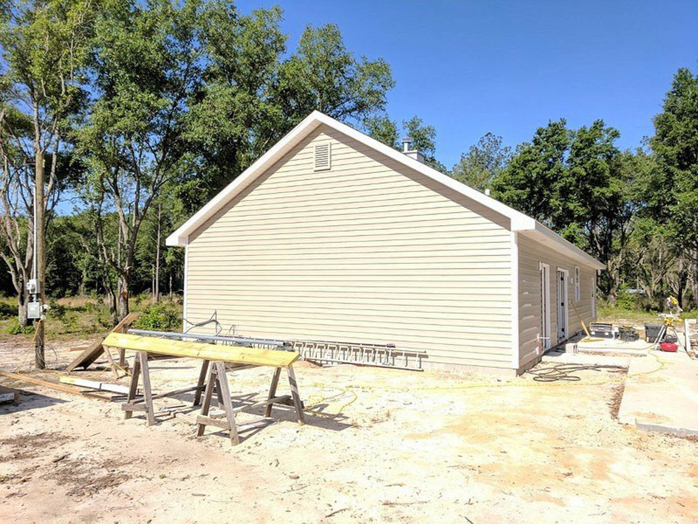 White house under construction with exposed wooden framing, roof installed, wooden sawhorse and benches on dirt ground, blue sky and trees in background, ladder leaning against