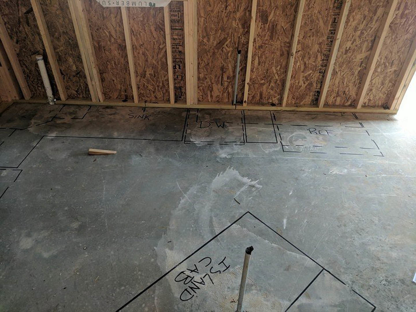 Concrete floor marked with black lines and text, surrounded by wood framing and a metal pole in an unfinished indoor space