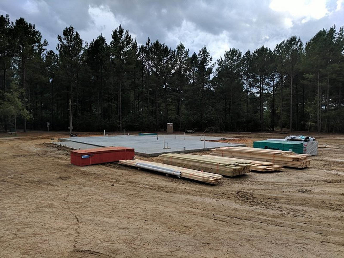 Framed wooden structure on a residential construction site, scattered lumber piles, red toolbox on dirt ground, parked car, metal pipe, surrounding trees under cloudy sky