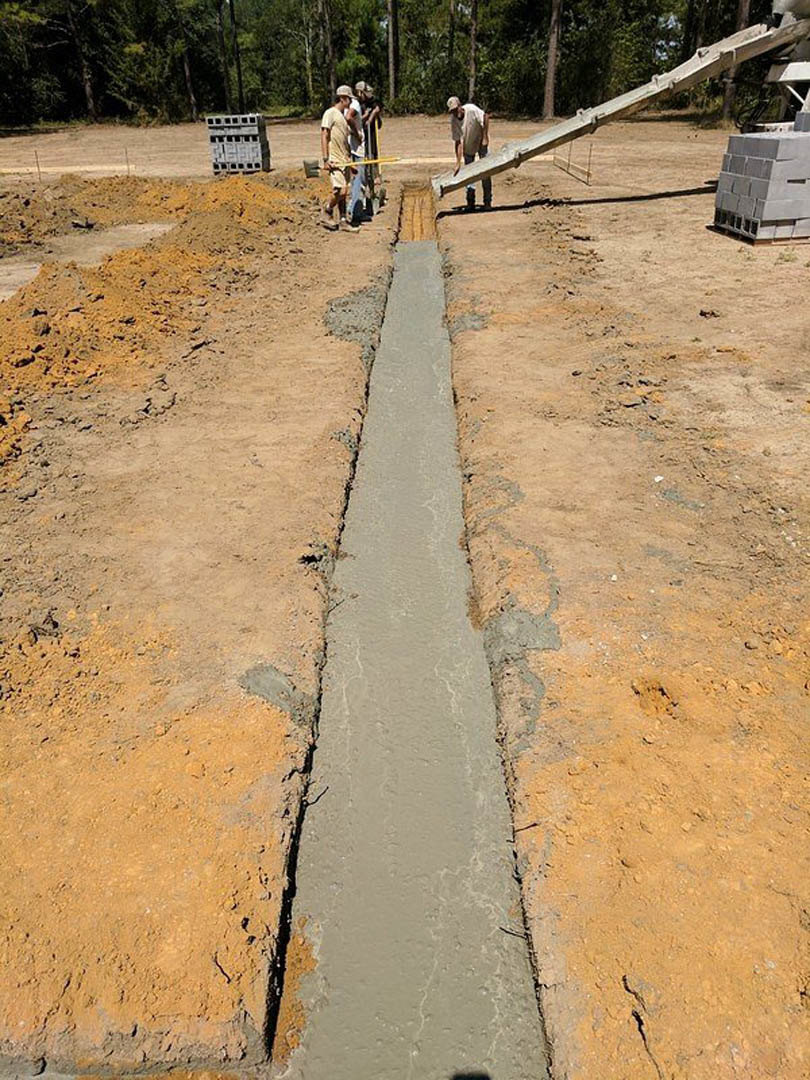 Two people working outdoors on a custom home construction site with exposed drainage ditch, stack of white bricks, scattered rocks, and trees in the background