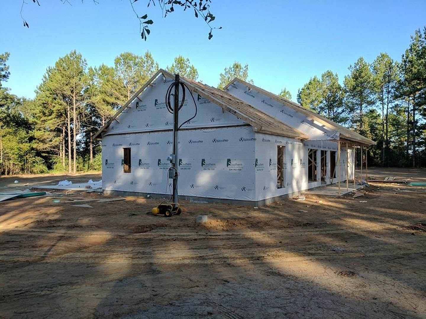 Framed house under construction with exposed wood roof, surrounded by trees and blue sky