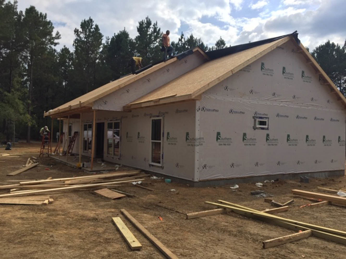 Framed house under construction with workers on the roof, scattered lumber on the ground, cloudy sky and trees in the background