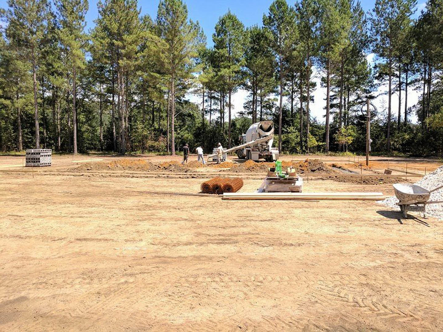 Cement mixer and wheelbarrow on residential construction site with white multi-windowed building, grassy ground, and trees in background