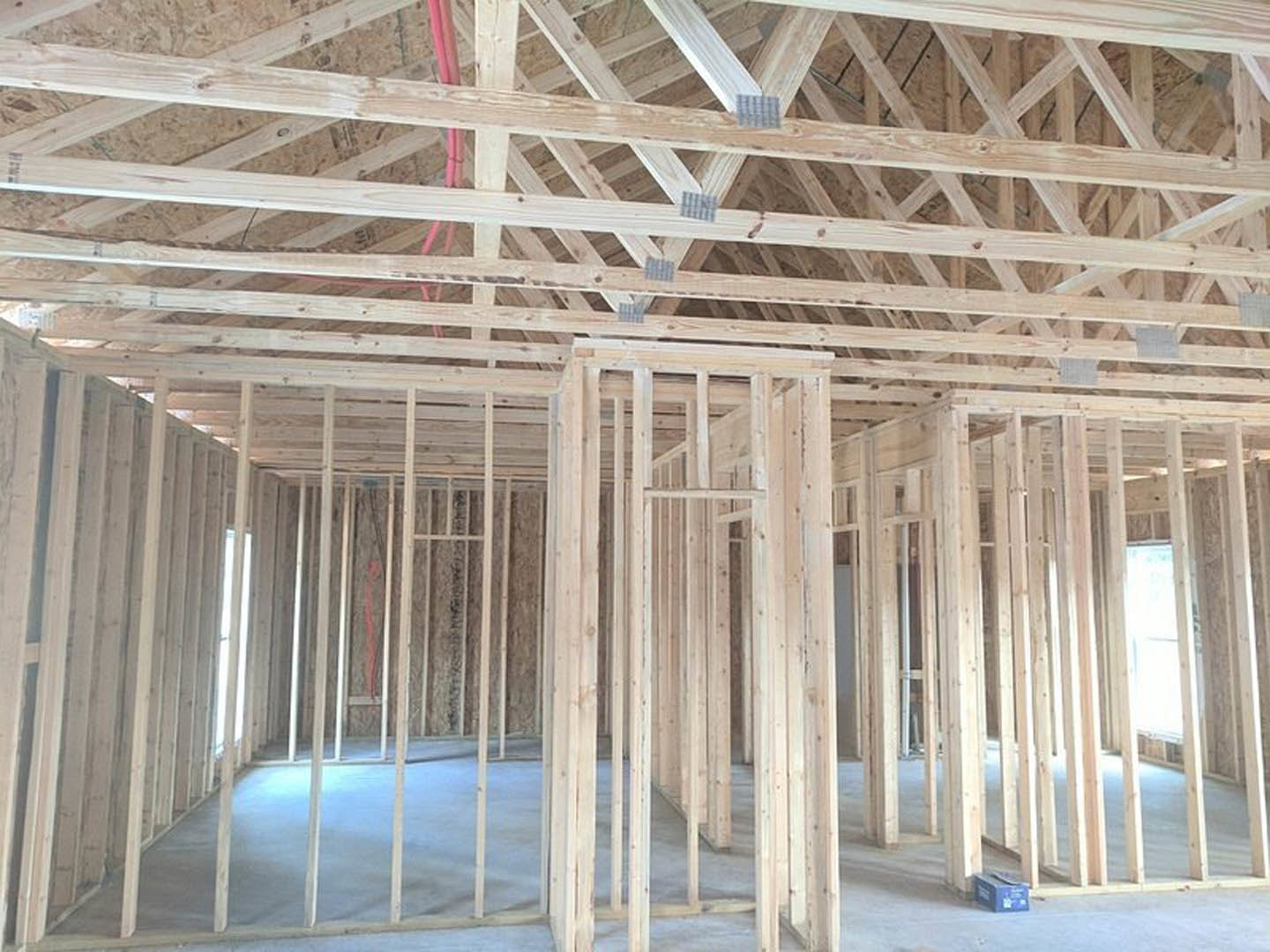 Wood framing and exposed beams inside a house under construction, with a wooden roof structure, red pipe, and window illuminated by natural light.